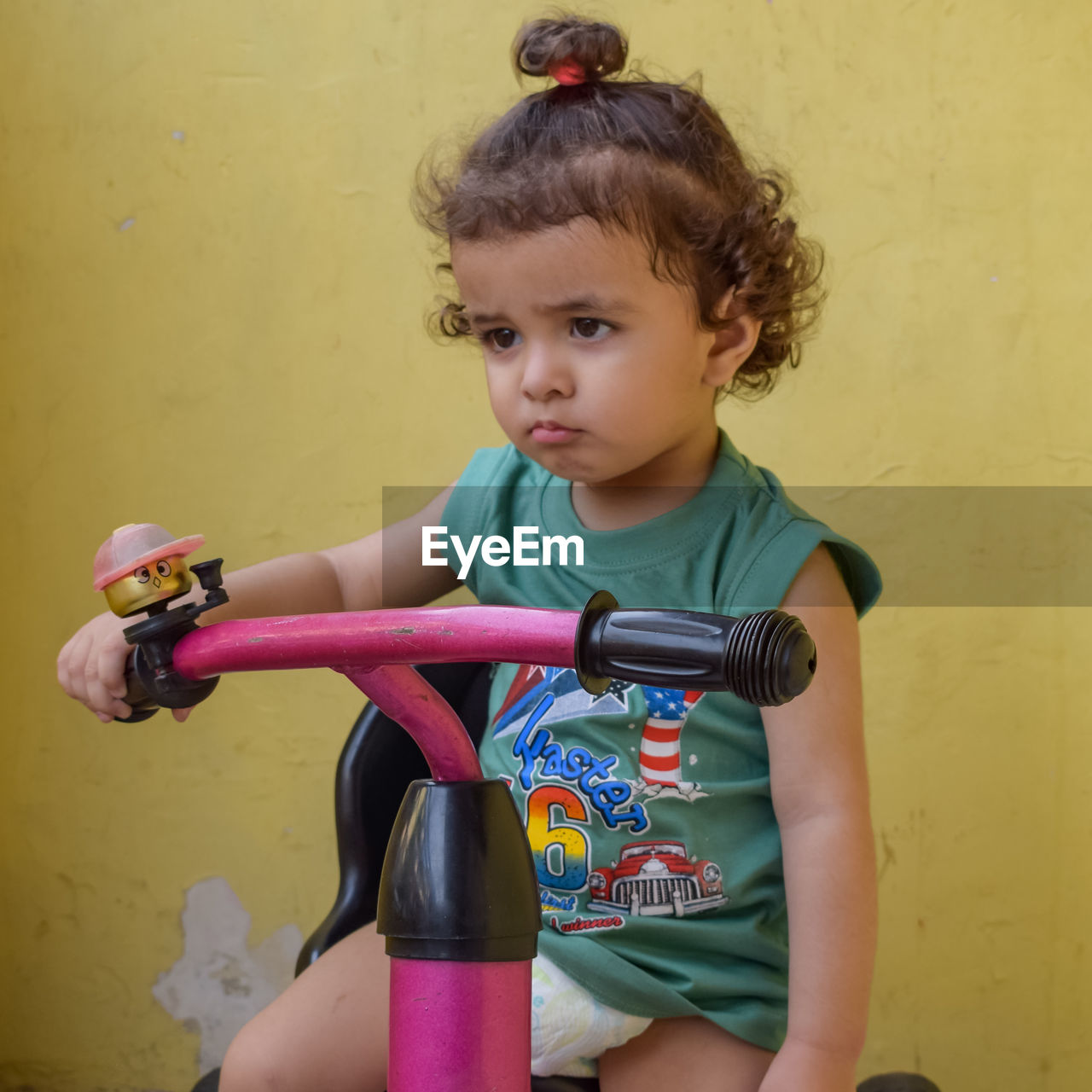 childhood, child, bicycle, one person, toddler, female, women, three quarter length, lifestyles, person, yellow, innocence, clothing, portrait, cute, sitting, holding, sports, wall - building feature, casual clothing, land vehicle, leisure activity, front view, indoors, baby, brown hair, toy, vehicle, looking at camera