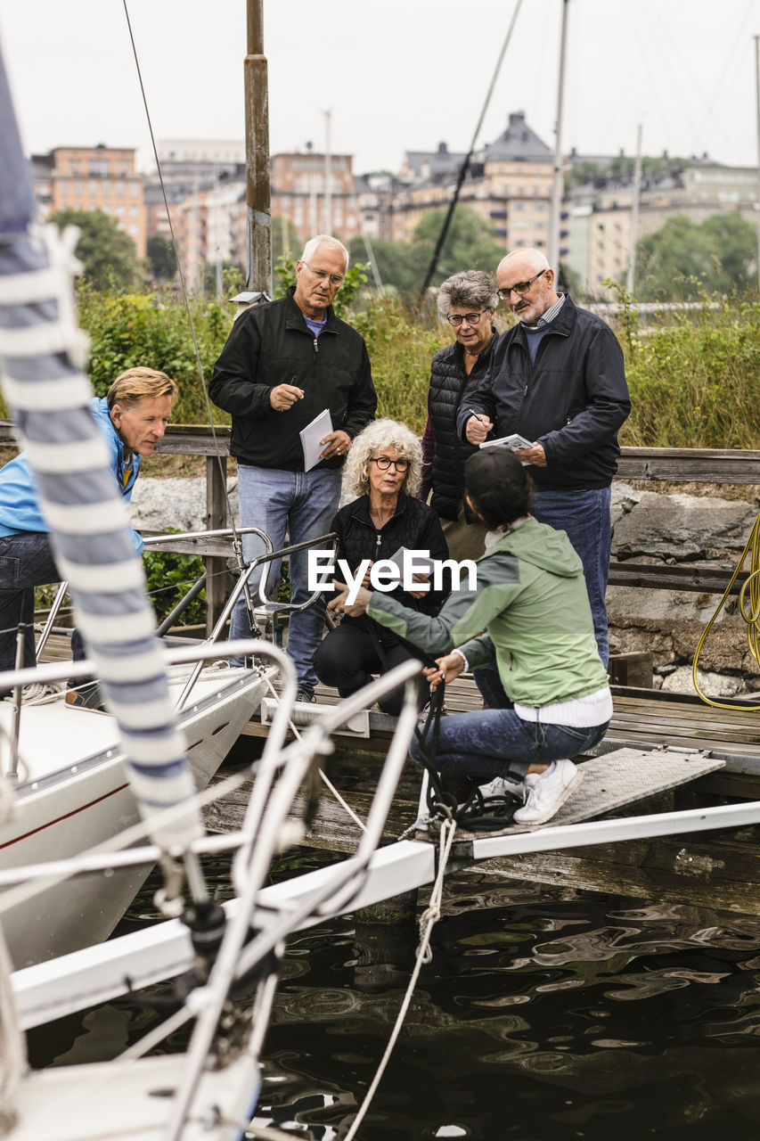 Senior men and women listening to female instructor by yacht during boat master course