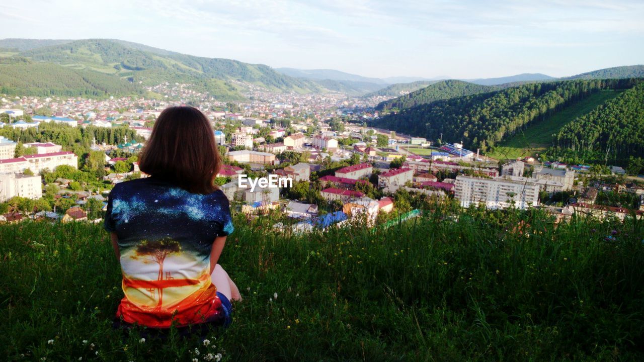 Rear view of woman relaxing on grassy hill against sky in city