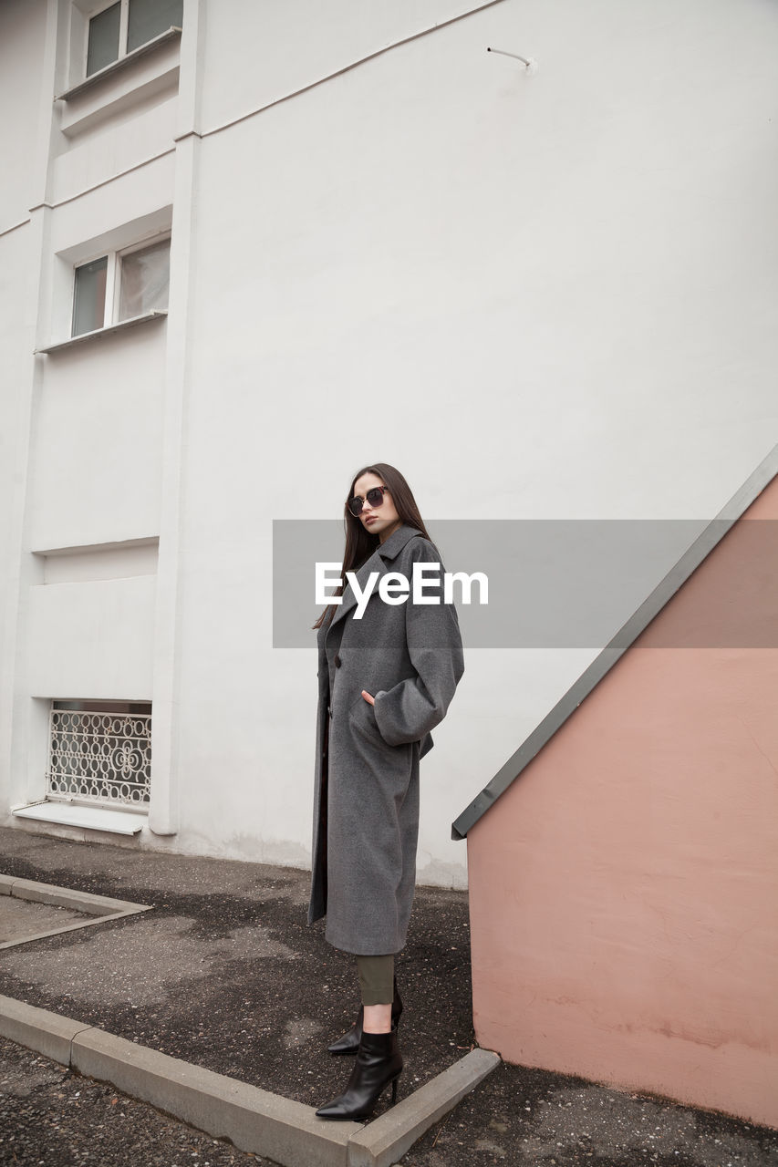 Full length side view portrait of young woman in grey coat and suit against wall outdoors, outwear 