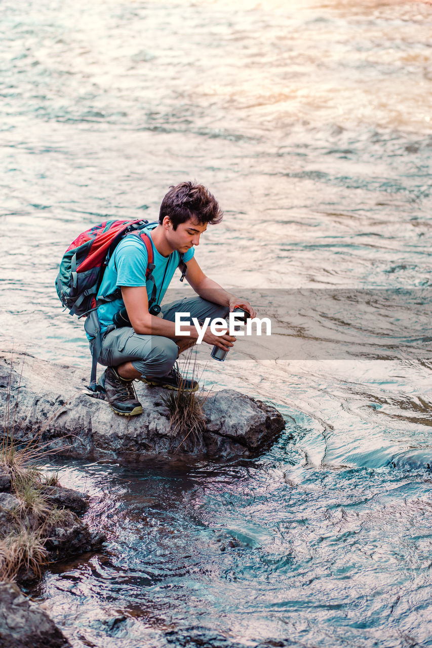 Boy with water bottle crouching at riverbank