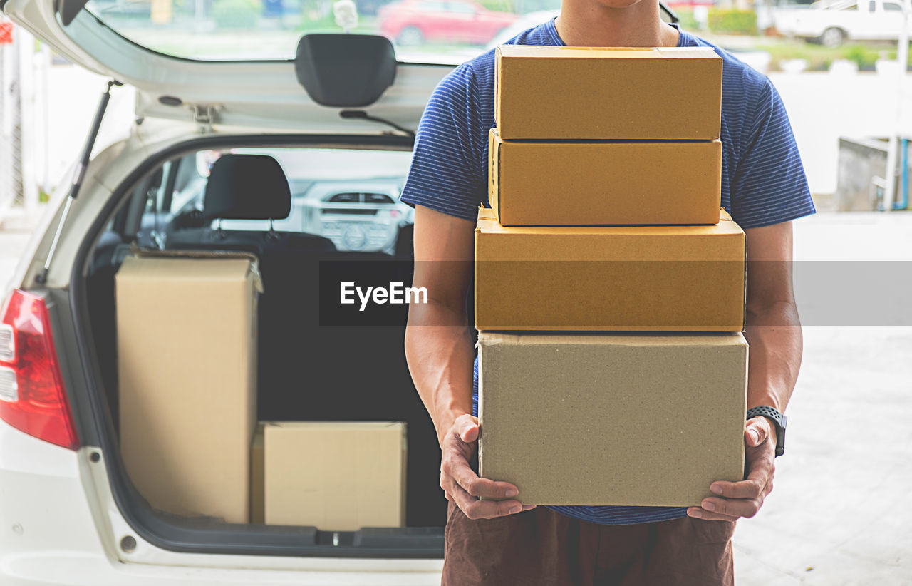 Midsection of delivery person holding stacked cardboard boxes against car