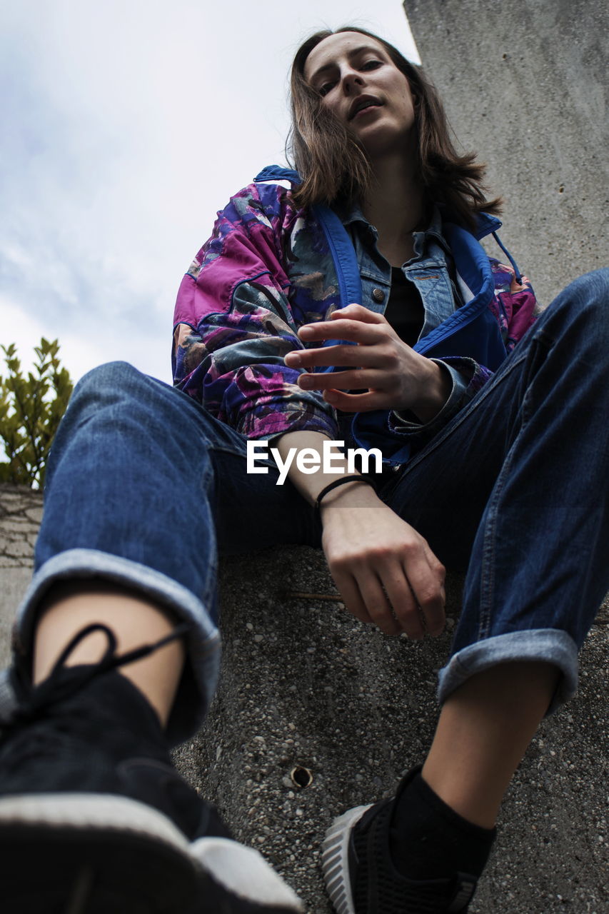 Low angle portrait of young woman sitting on retaining wall against sky