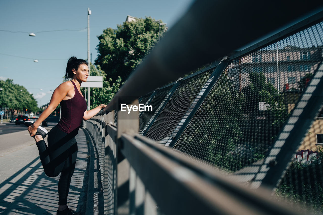 Young sportswoman stretching leg by railing on bridge in city