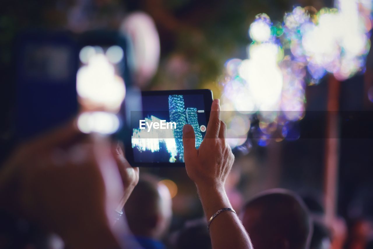 Cropped hands photographing illuminated lights with digital tablet at night