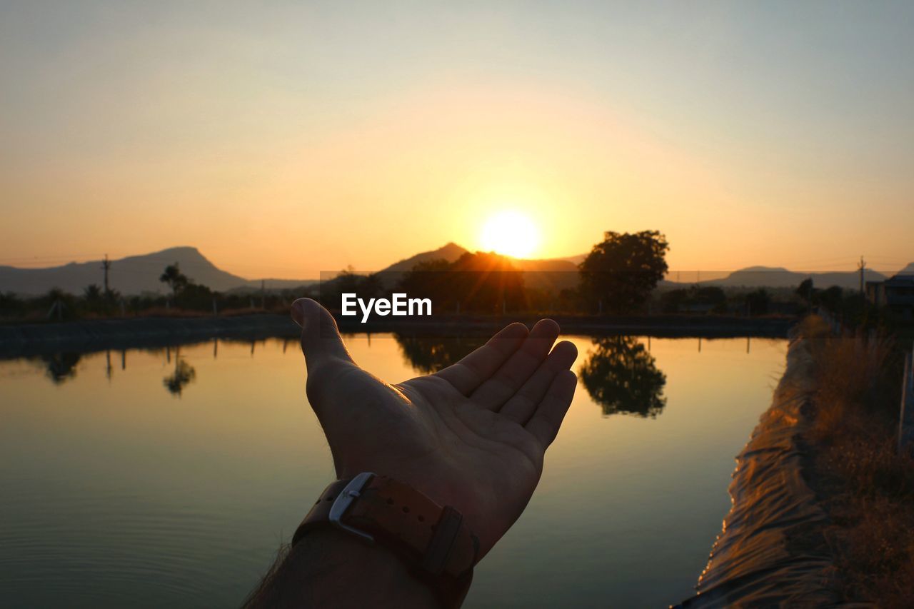 Cropped hand gesturing towards lake during sunset