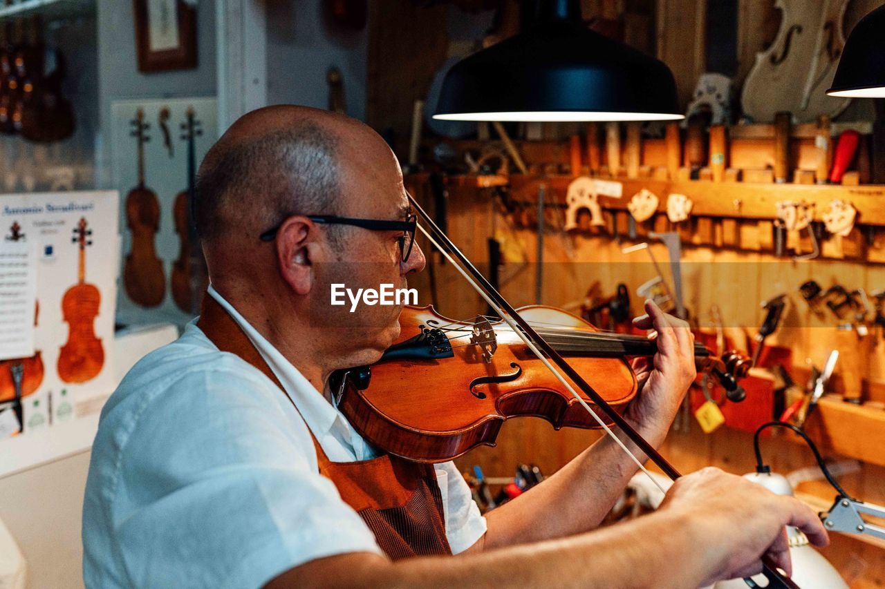 Side view of mature male artisan luthier testing violin after restoration in workshop