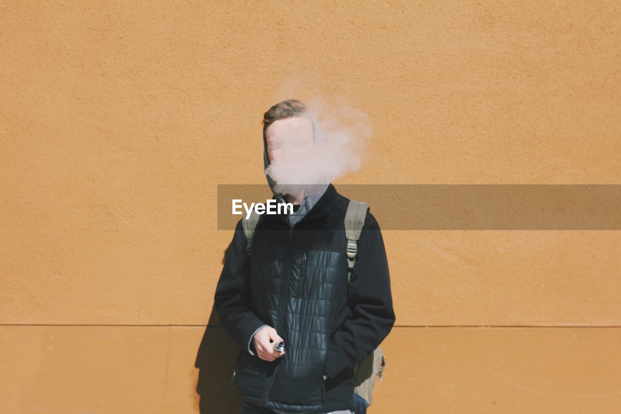 Young man smoking while standing against wall on sunny day