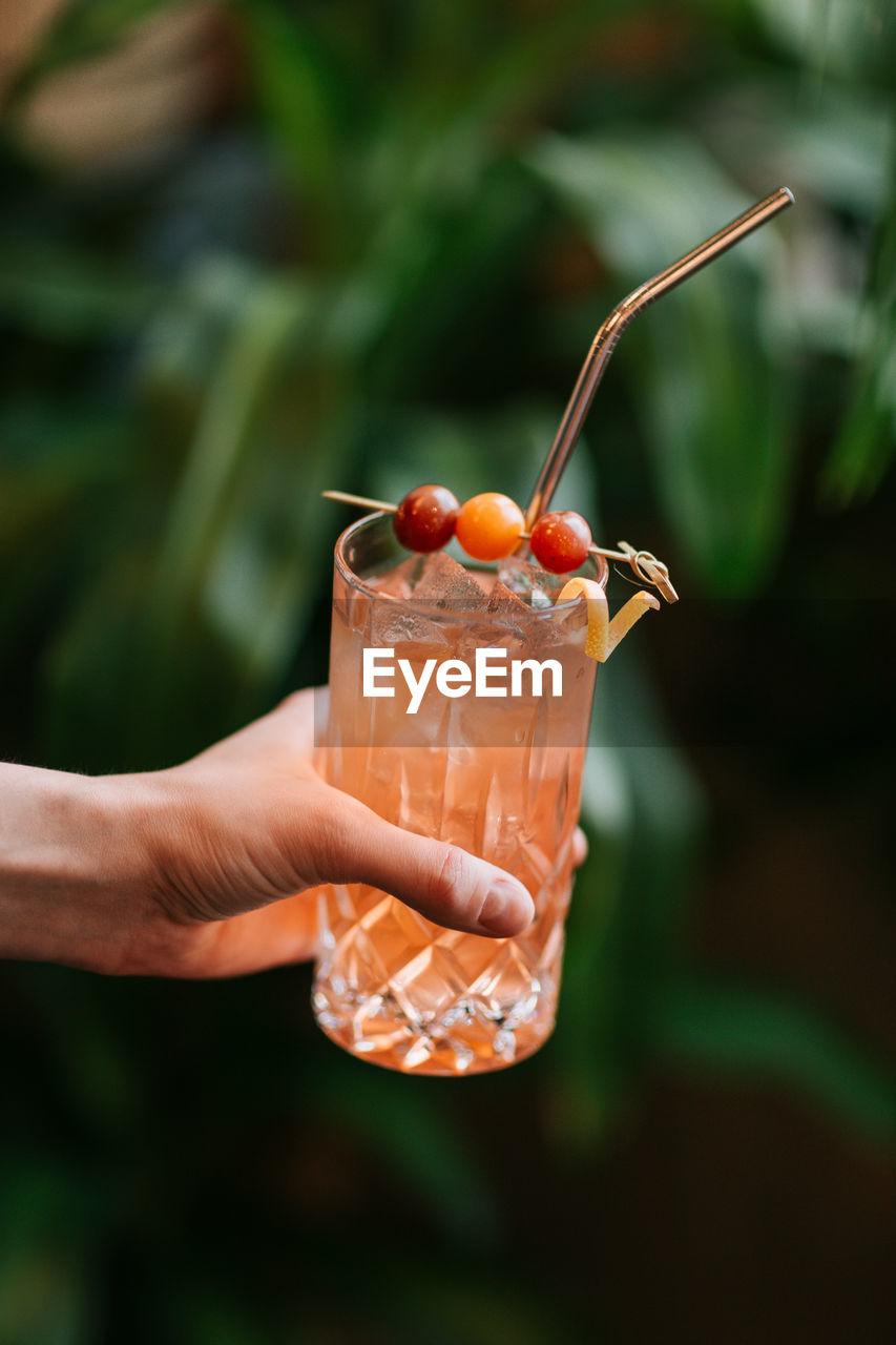 Crop anonymous person demonstrating transparent glass with cold cocktail with ice cubes served with berries on stick and zero waste straw person