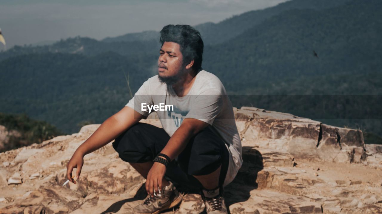 Young man looking away and smoking while sitting on rock against mountains