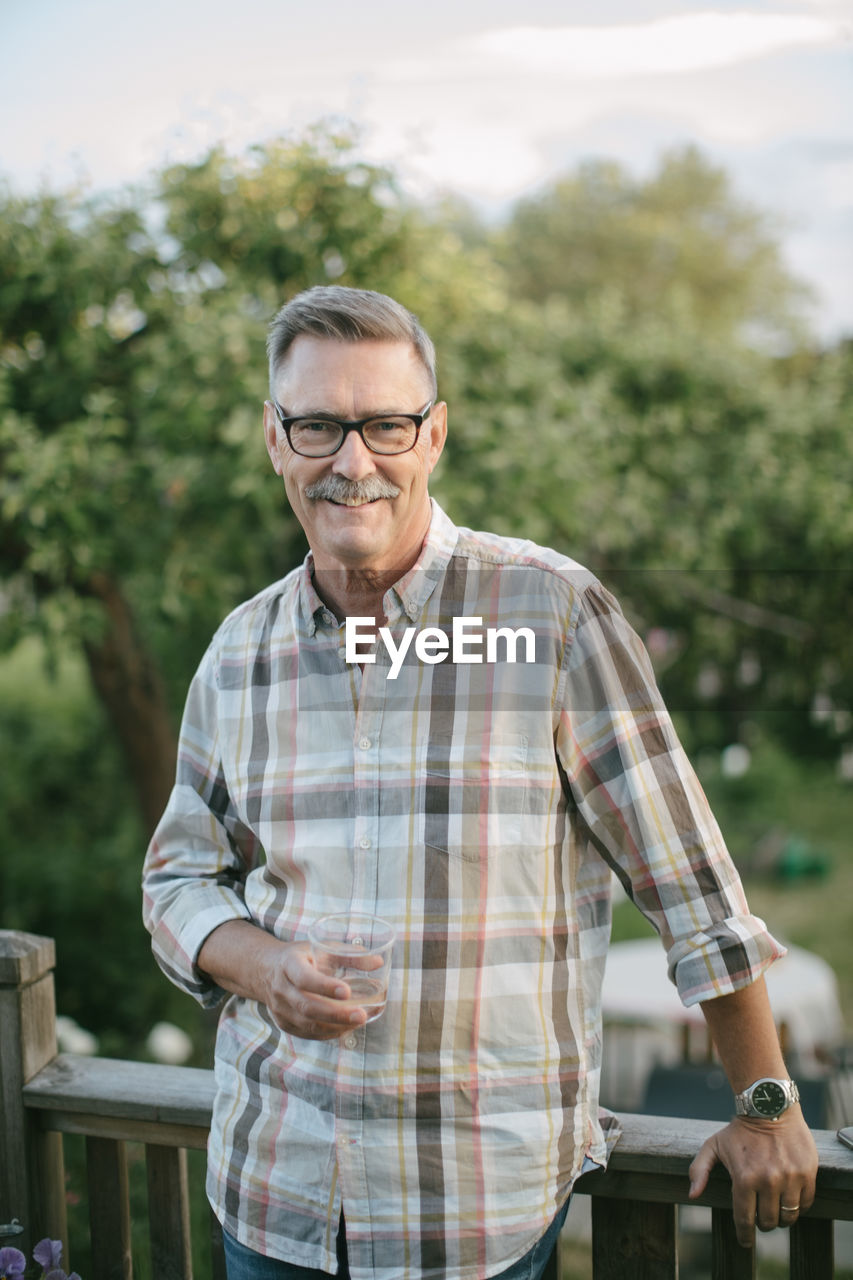 Portrait of smiling senior man holding drinking glass standing on porch