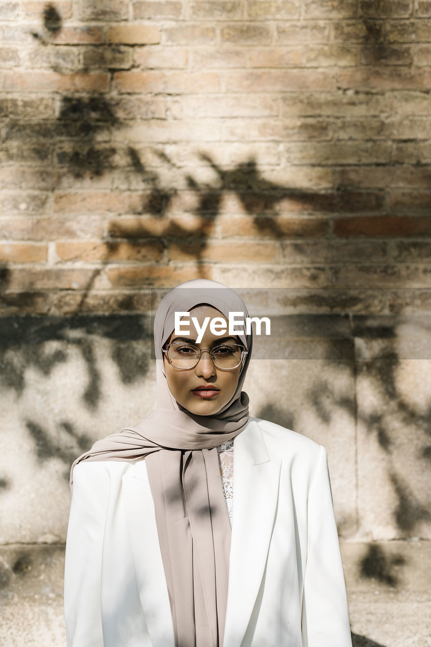 Woman wearing eyeglasses while standing in front of wall on sunny day