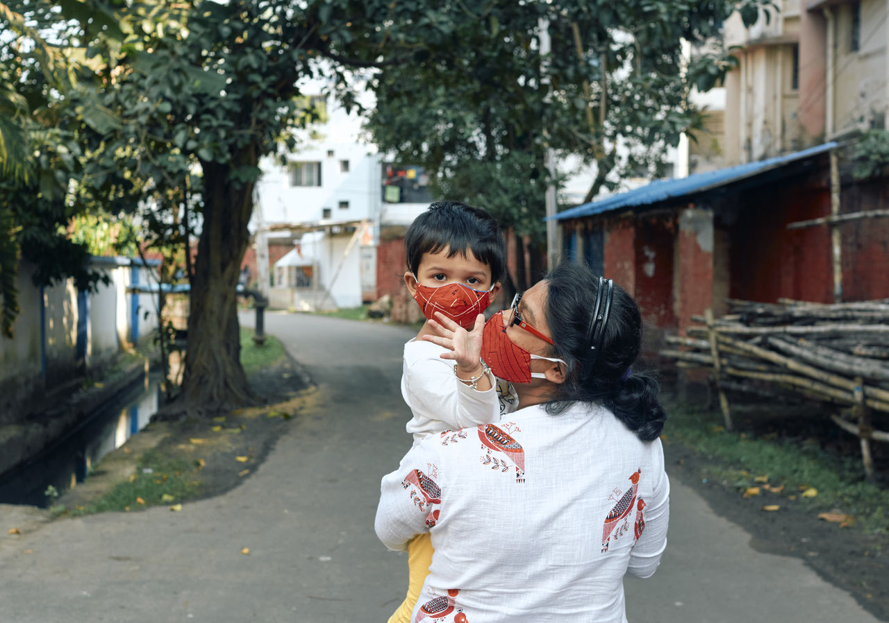 A cute baby girl in her mother's arm, walking in street. they are wearing protective face masks.