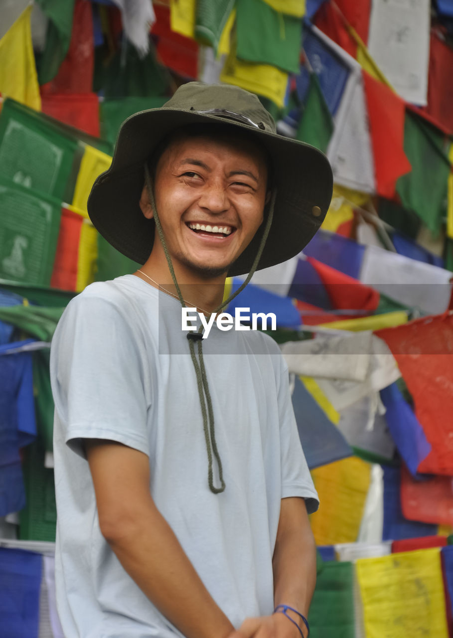 A happy tibetan young man looking sideways, standing against buddhist prayer flags 