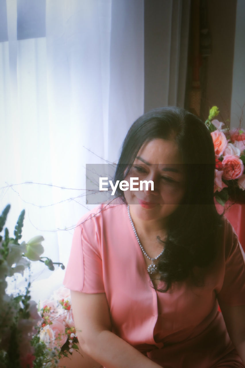 one person, flower, flowering plant, plant, women, adult, female, pink, young adult, nature, indoors, front view, looking, looking down, waist up, sitting, emotion, lifestyles, flower arrangement, day, beauty in nature, bouquet, brown hair, hairstyle, contemplation, rose, person, floristry, bride, red, portrait, window, black hair, long hair, dress, casual clothing, smiling, sunlight, leisure activity, eyes closed, clothing