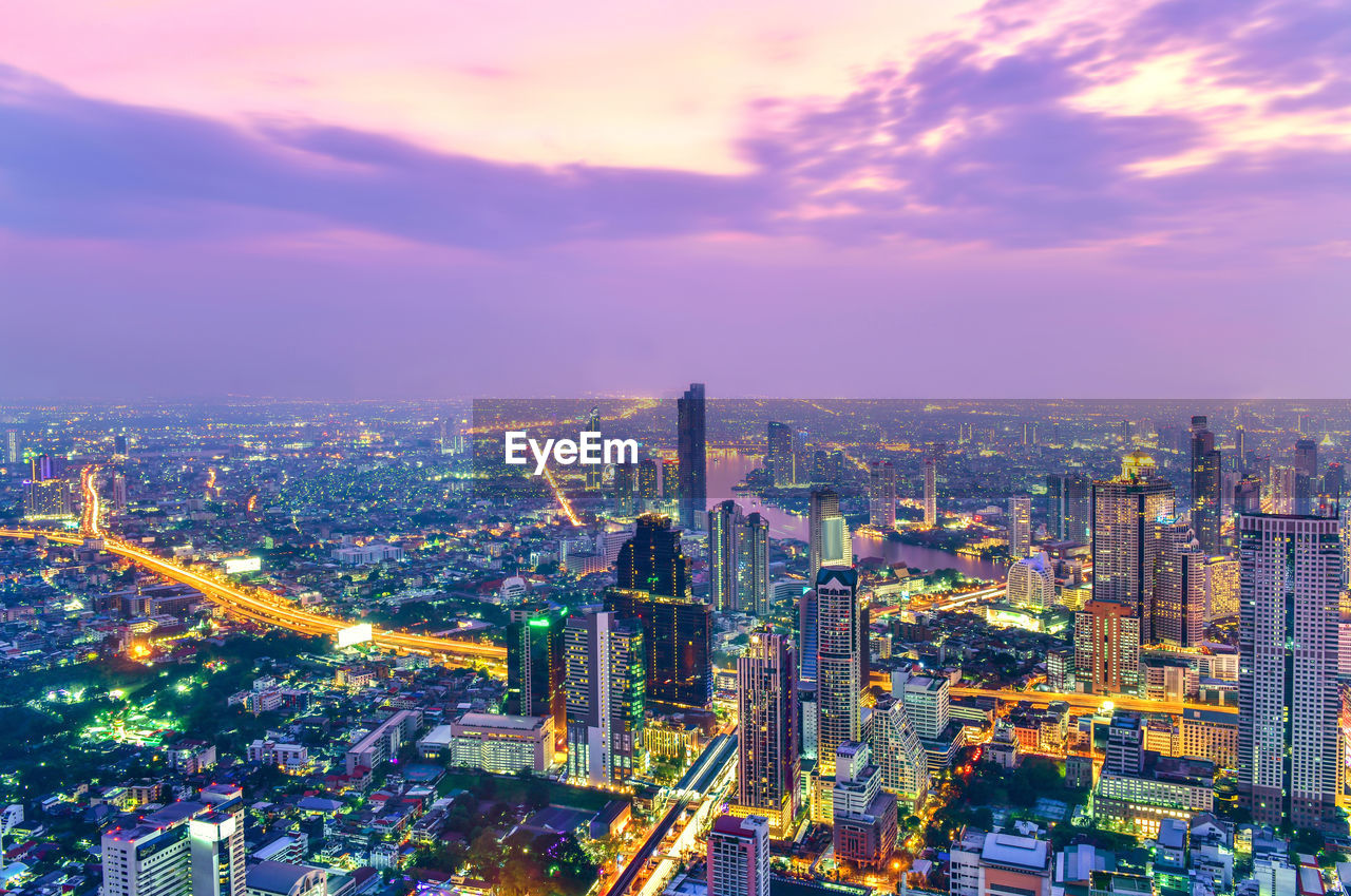 HIGH ANGLE VIEW OF ILLUMINATED CITYSCAPE AGAINST SKY AT SUNSET