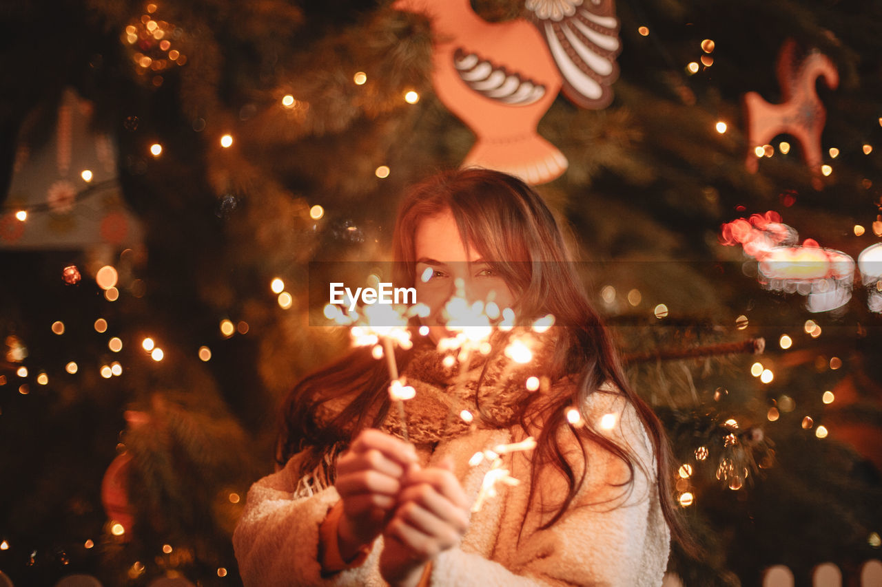 Happy teenage girl holding sparklers standing by christmas tree