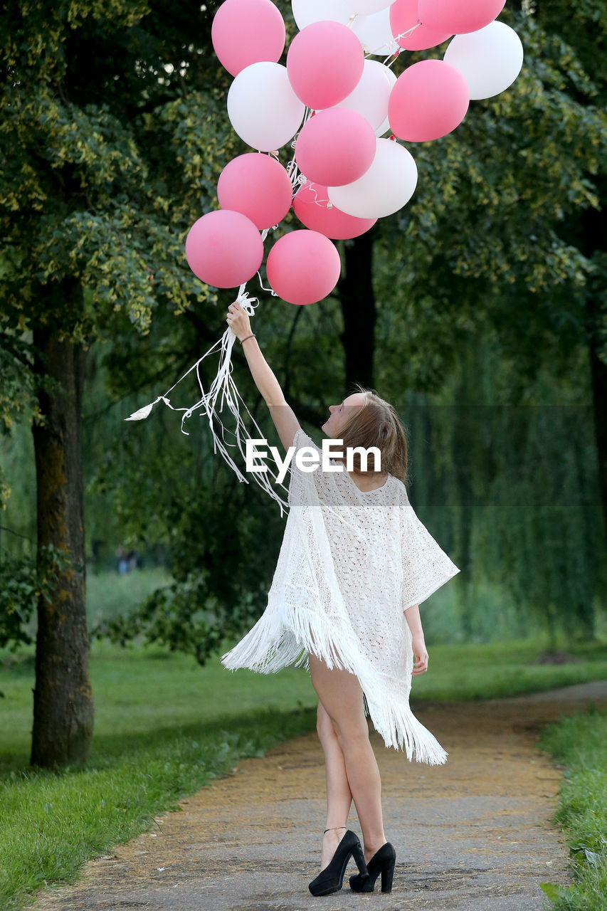 Woman holding pink and white helium balloons on footpath against trees at park