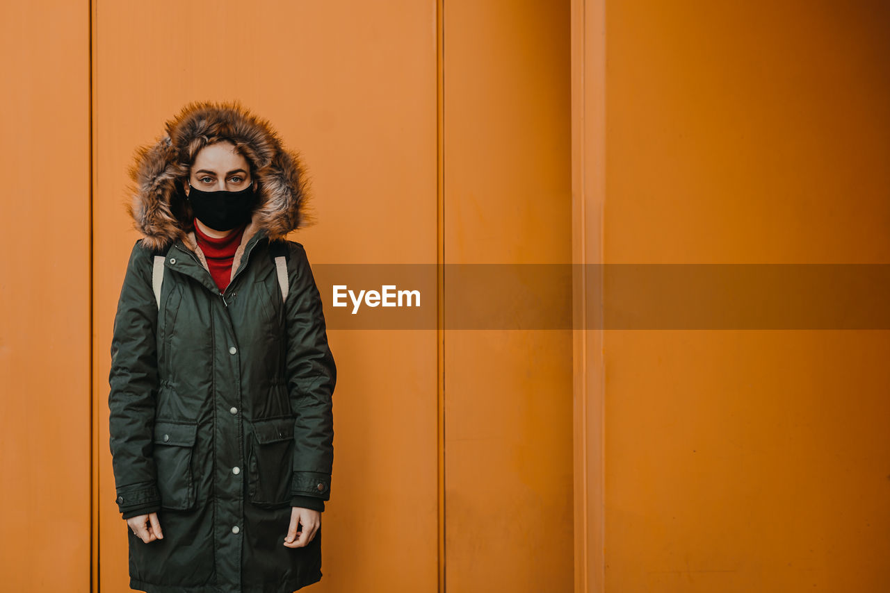 Portrait of young woman wearing mask standing against yellow wall