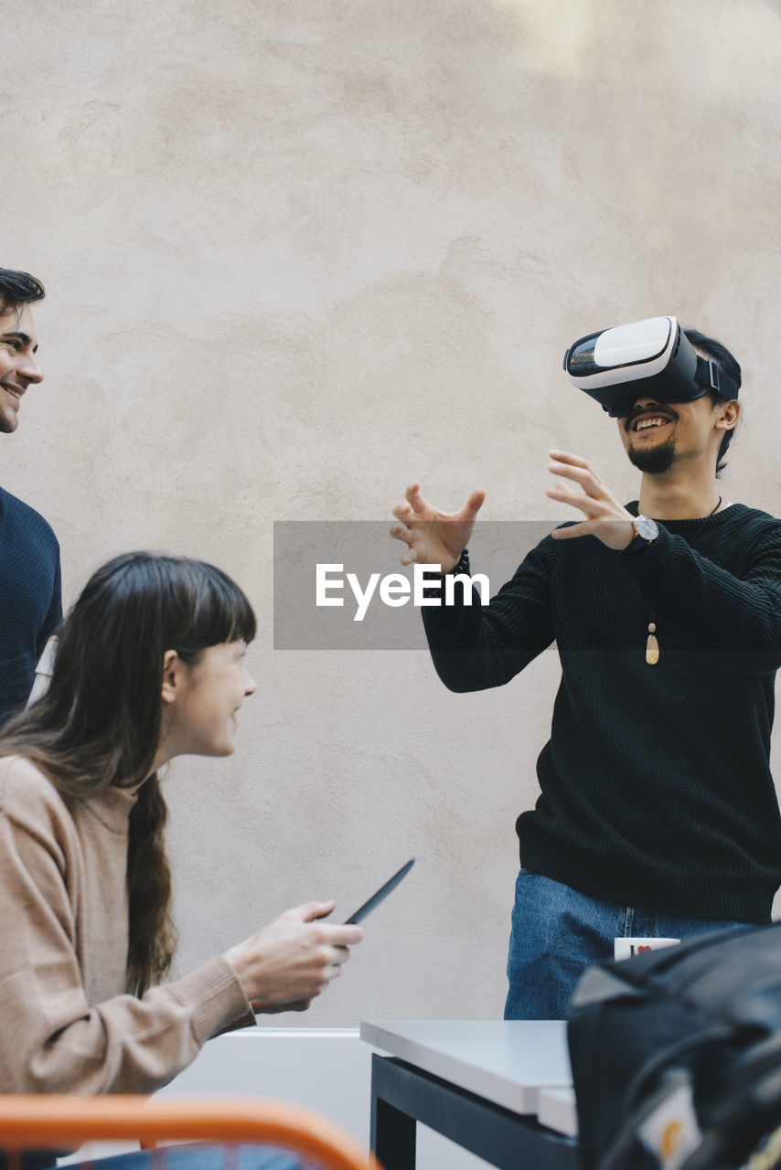 Smiling male computer programmer gesturing while using vr glasses by colleagues in office