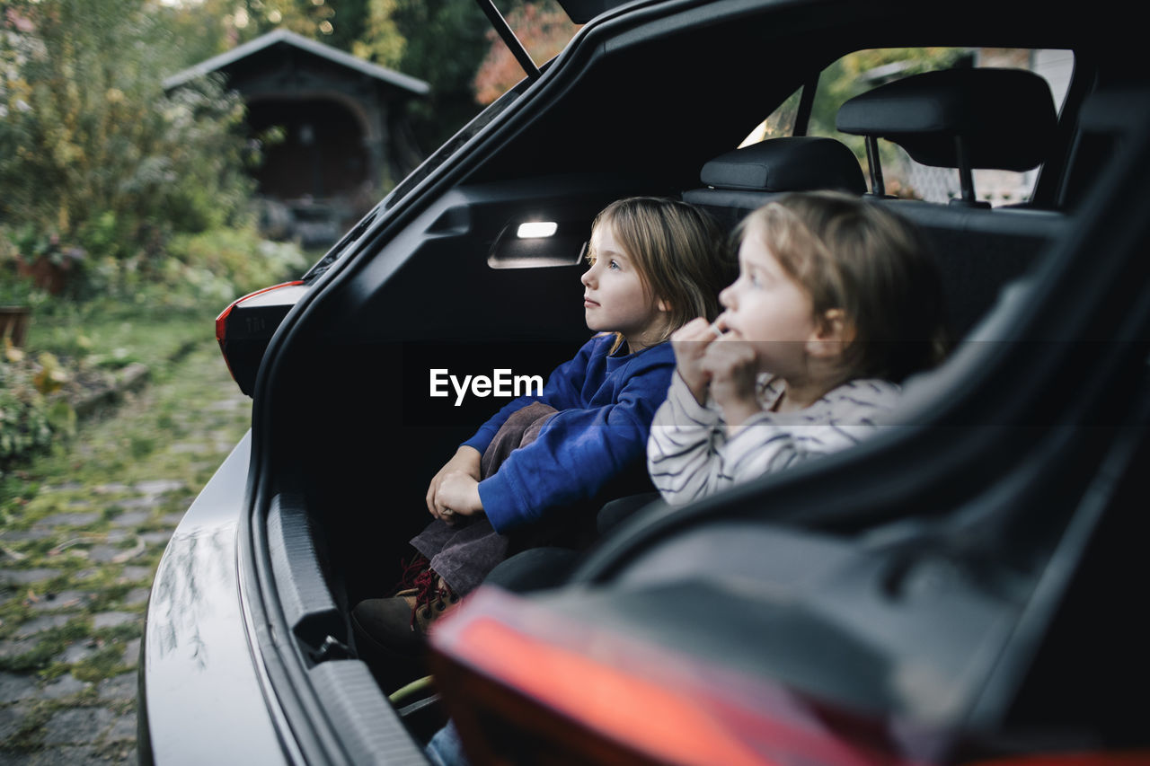 Girl day dreaming by sister while sitting in electric car trunk