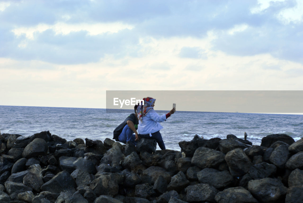 WOMAN SITTING ON ROCKS AT BEACH AGAINST SKY