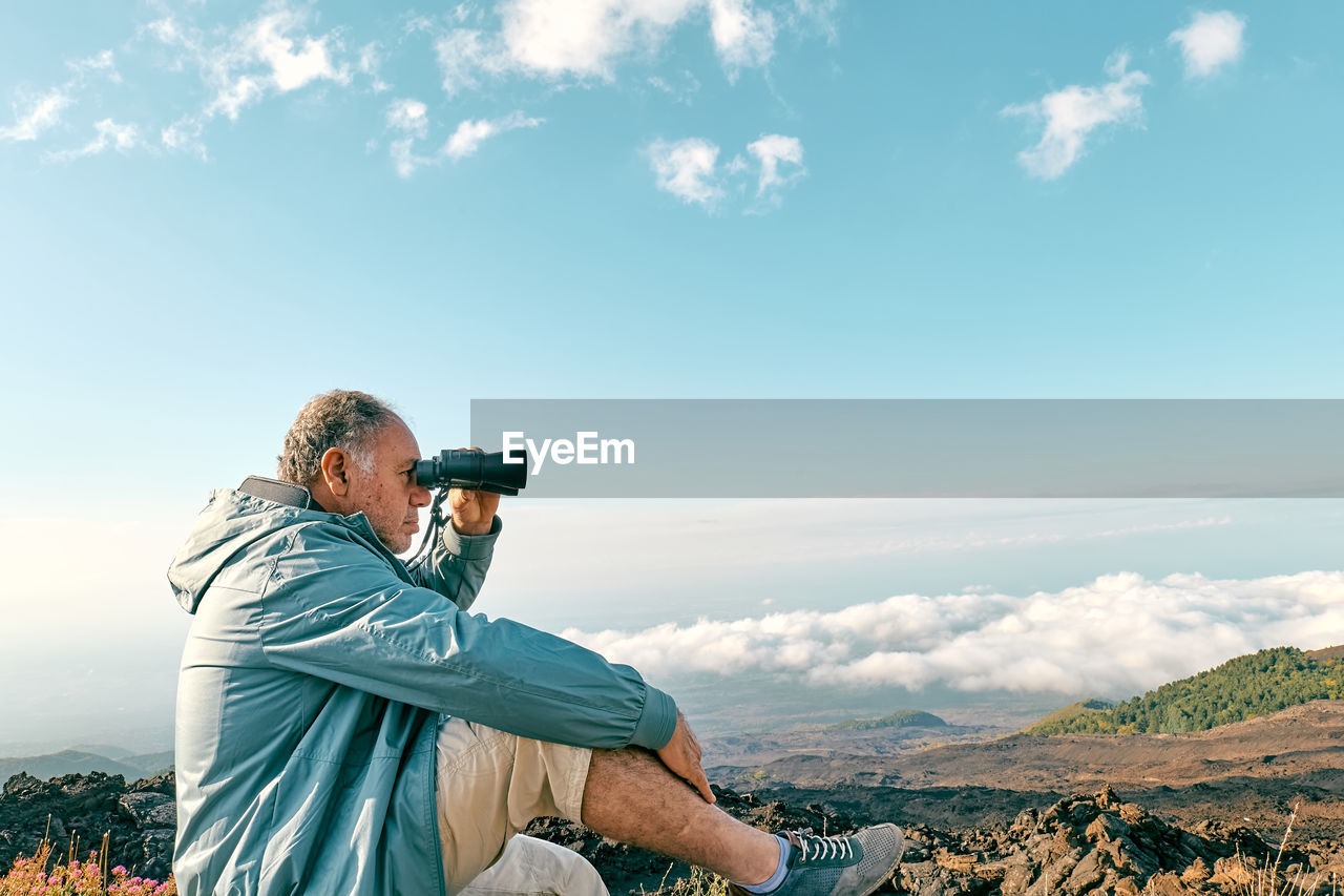 Rear view of man looking through binoculars at panoramic view of summits of active volcano etna