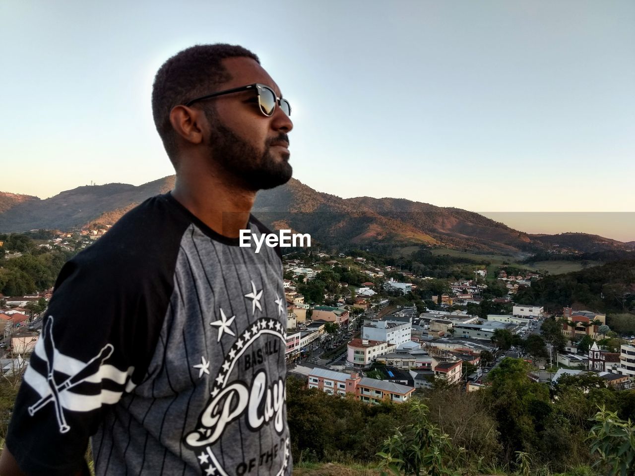YOUNG MAN WEARING SUNGLASSES STANDING AGAINST MOUNTAIN IN CITY
