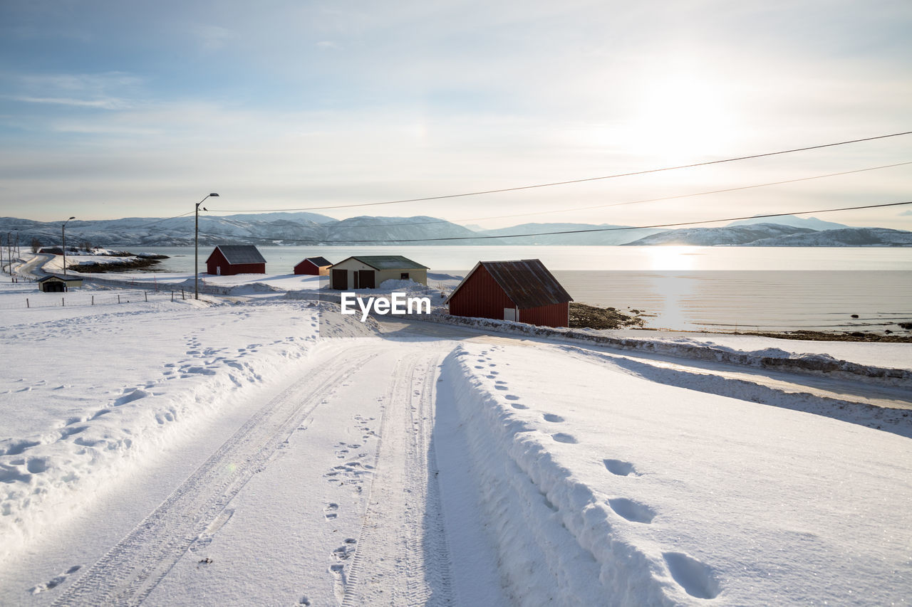 Snow covered road leading towards houses at lakeshore against sky