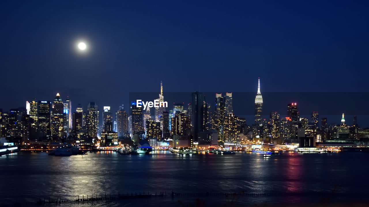 Illuminated cityscape by river against clear sky at night