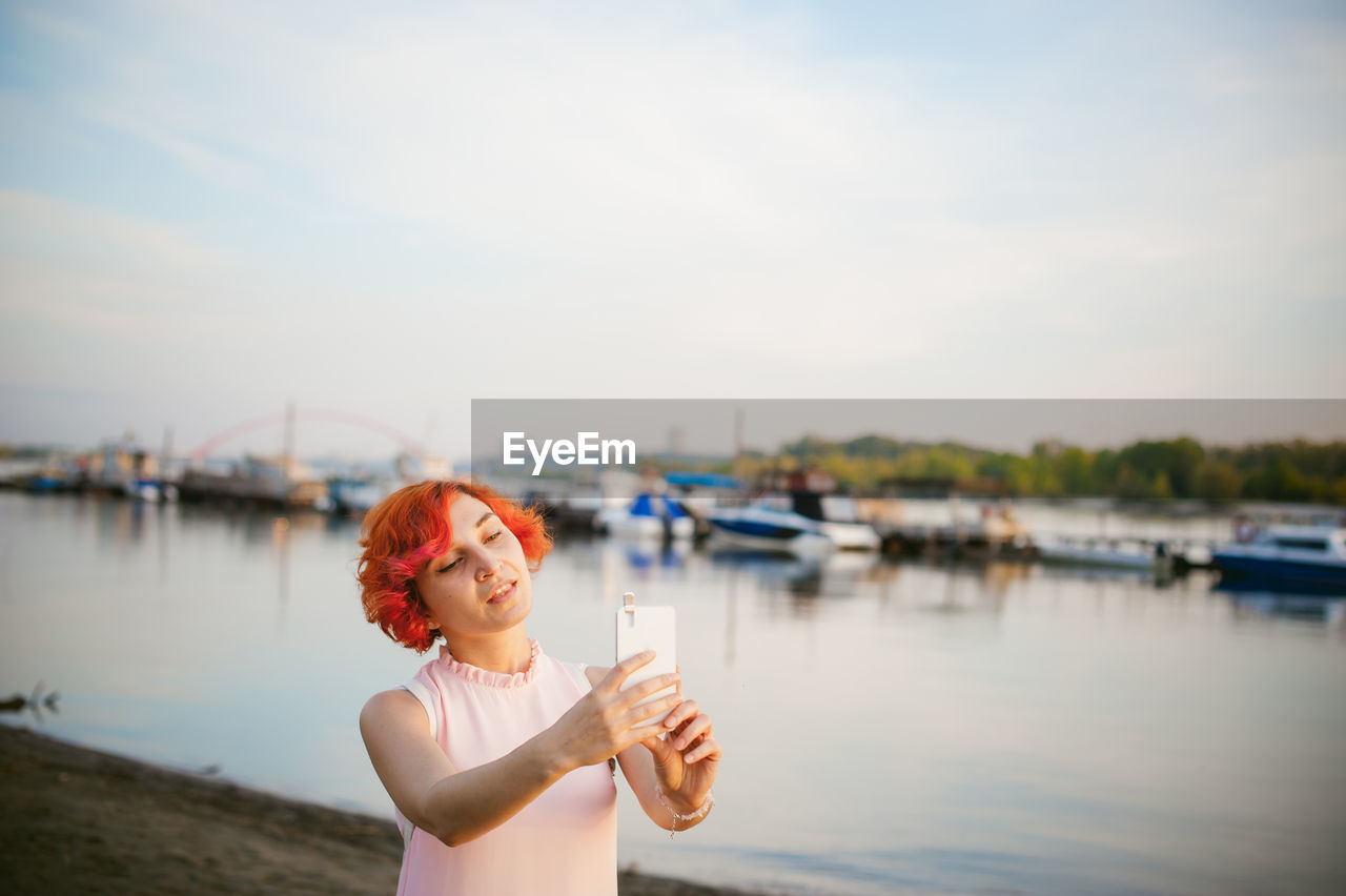 Mid adult woman taking selfie with mobile phone while standing by river against sky