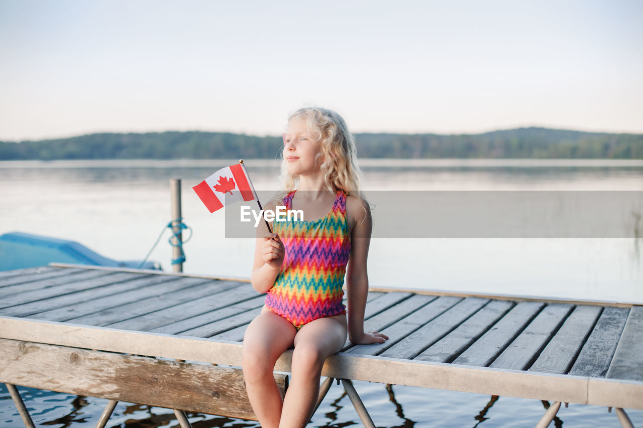 Happy caucasian girl sitting on pier by lake and waving canadian flag. kid celebrating canada day 