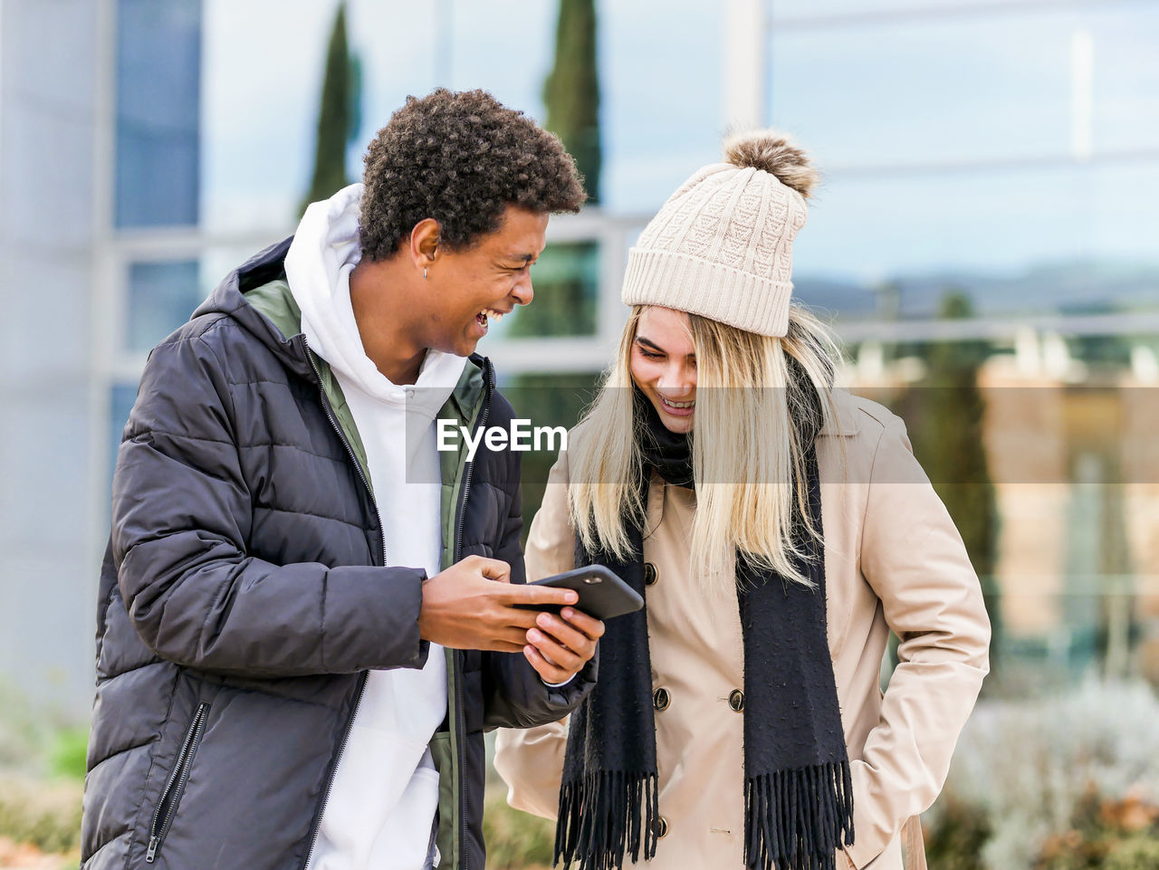 Young sincere multiethnic partners in warm clothes watching mobile phone while spending time together in town