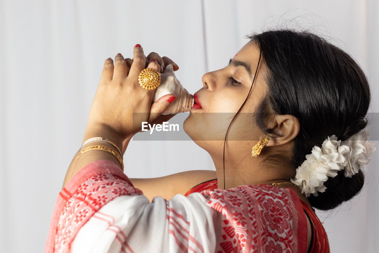 A beautiful indian woman in red saree blowing conch shell on white background