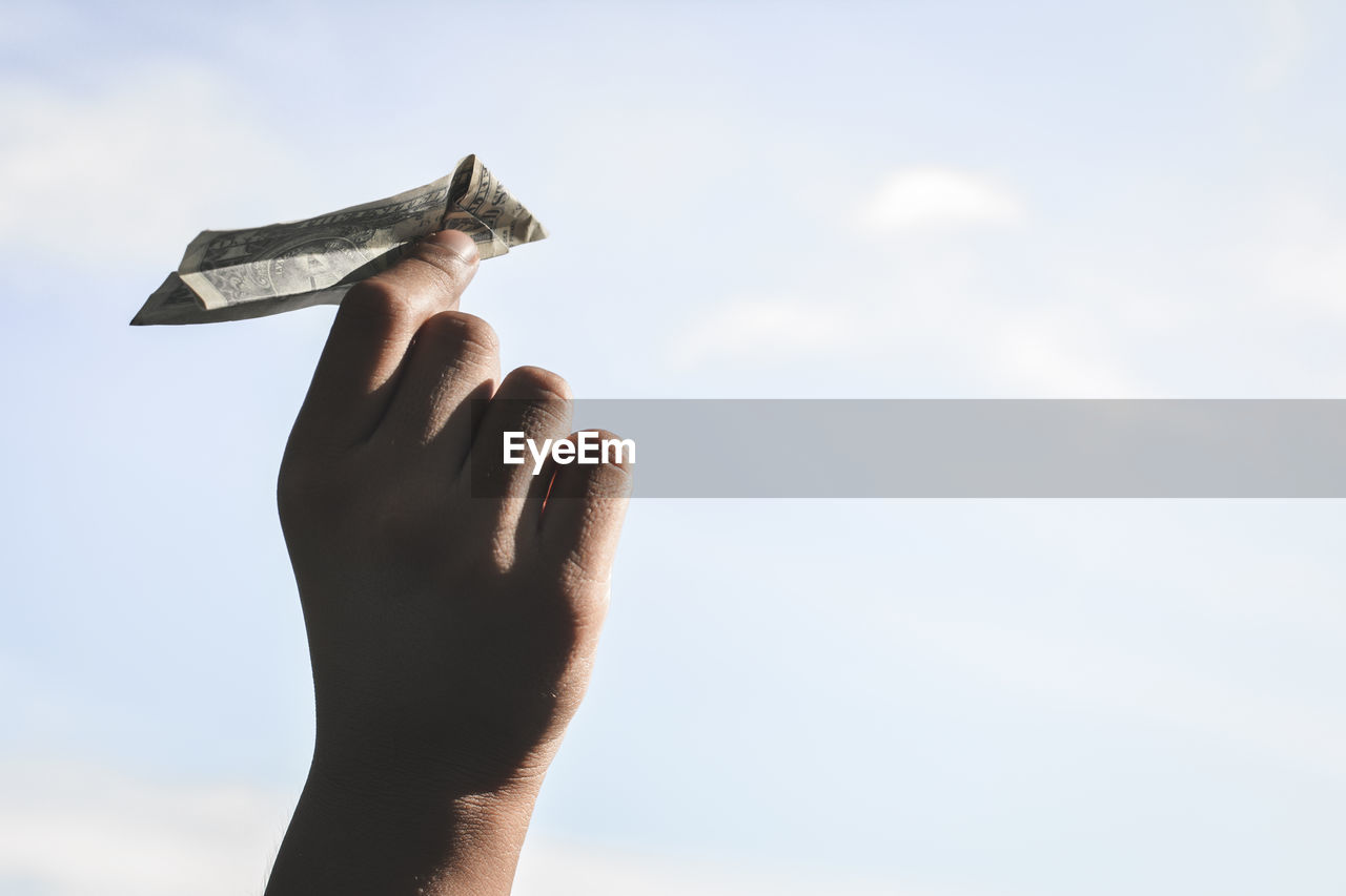 Cropped image of hand holding paper currency airplane against sky