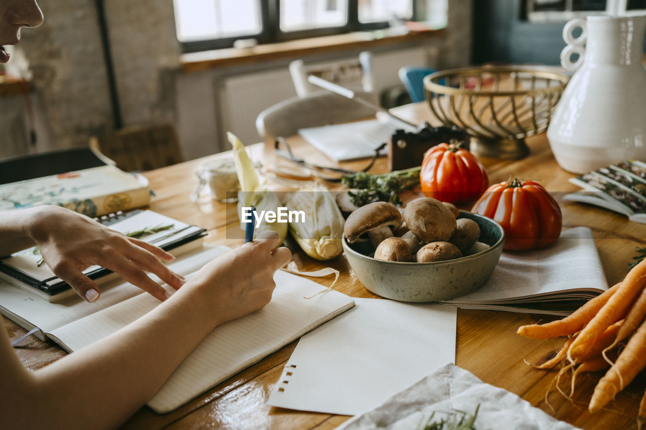 Hand of female food stylist writing in diary by fresh vegetables on table in studio