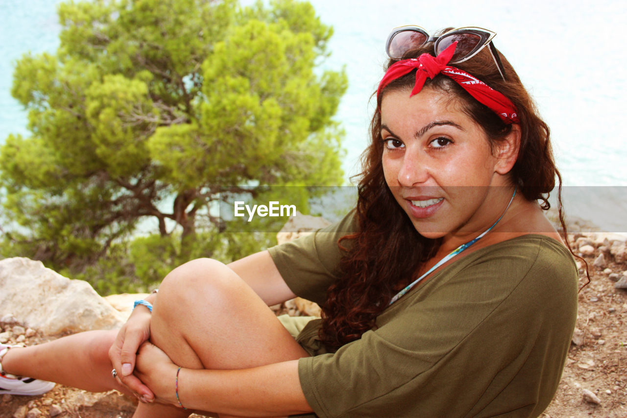 Caucasian girl smiles shyly sitting on a cliff with behind a blurred tree in summer 