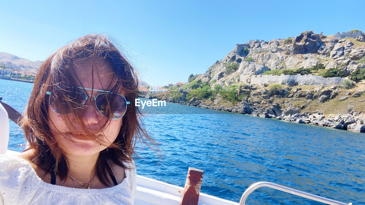 vacation, water, one person, women, sea, portrait, nature, sunglasses, adult, glasses, leisure activity, trip, holiday, headshot, travel, fashion, day, sunlight, sky, young adult, lifestyles, hairstyle, mountain, front view, sunny, female, summer, long hair, beauty in nature, outdoors, travel destinations, blue, transportation, smiling, brown hair, land, beach, clear sky, happiness, relaxation, nautical vessel, scenics - nature, clothing, tourism, enjoyment, person, emotion, ocean, casual clothing, boat, vehicle