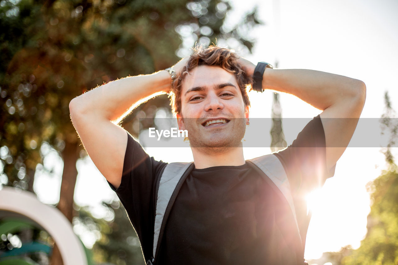 Portrait of young happy man with arms raised standing against sky