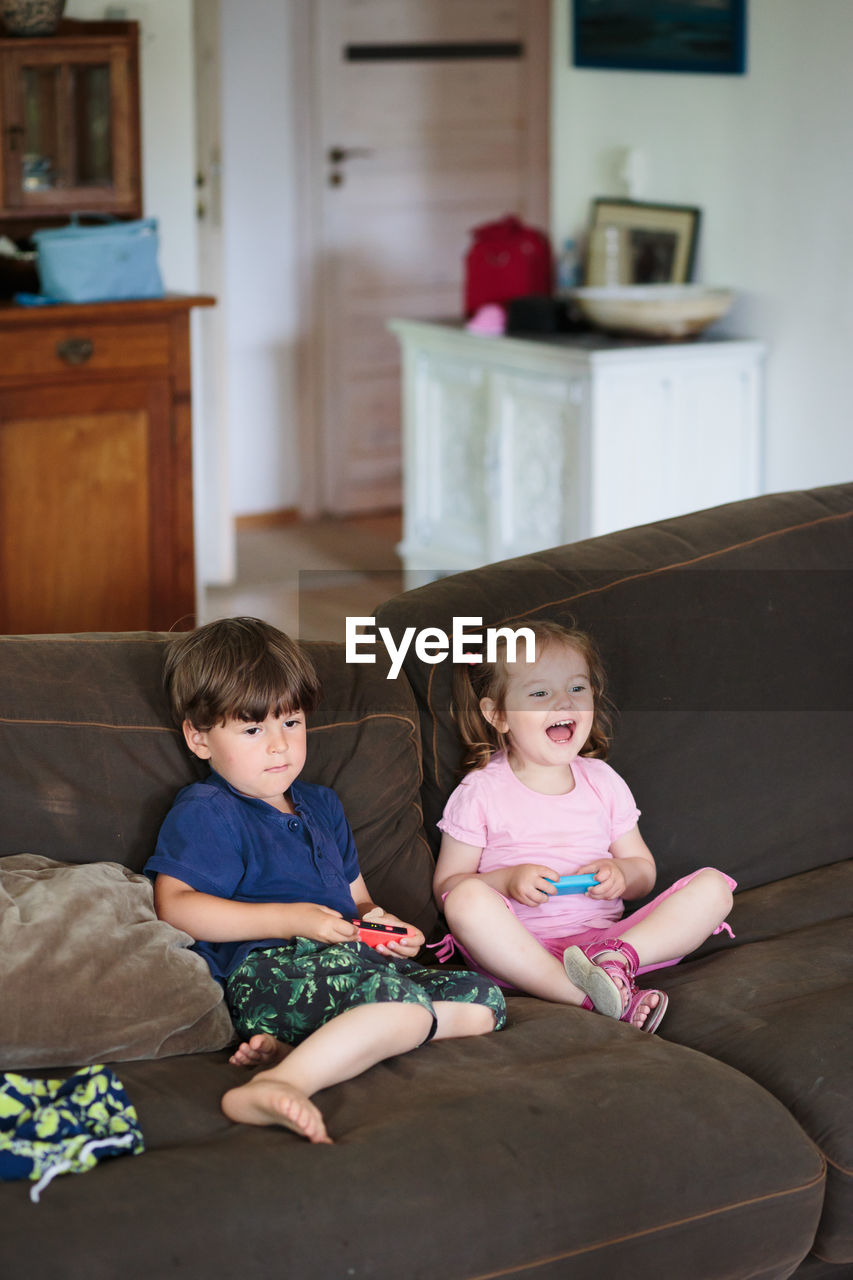 Siblings playing video game while sitting on sofa at home