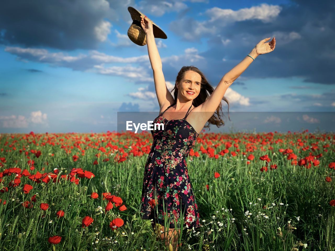 Portrait of happy young woman wearing dress and straw hat in a field of poppies