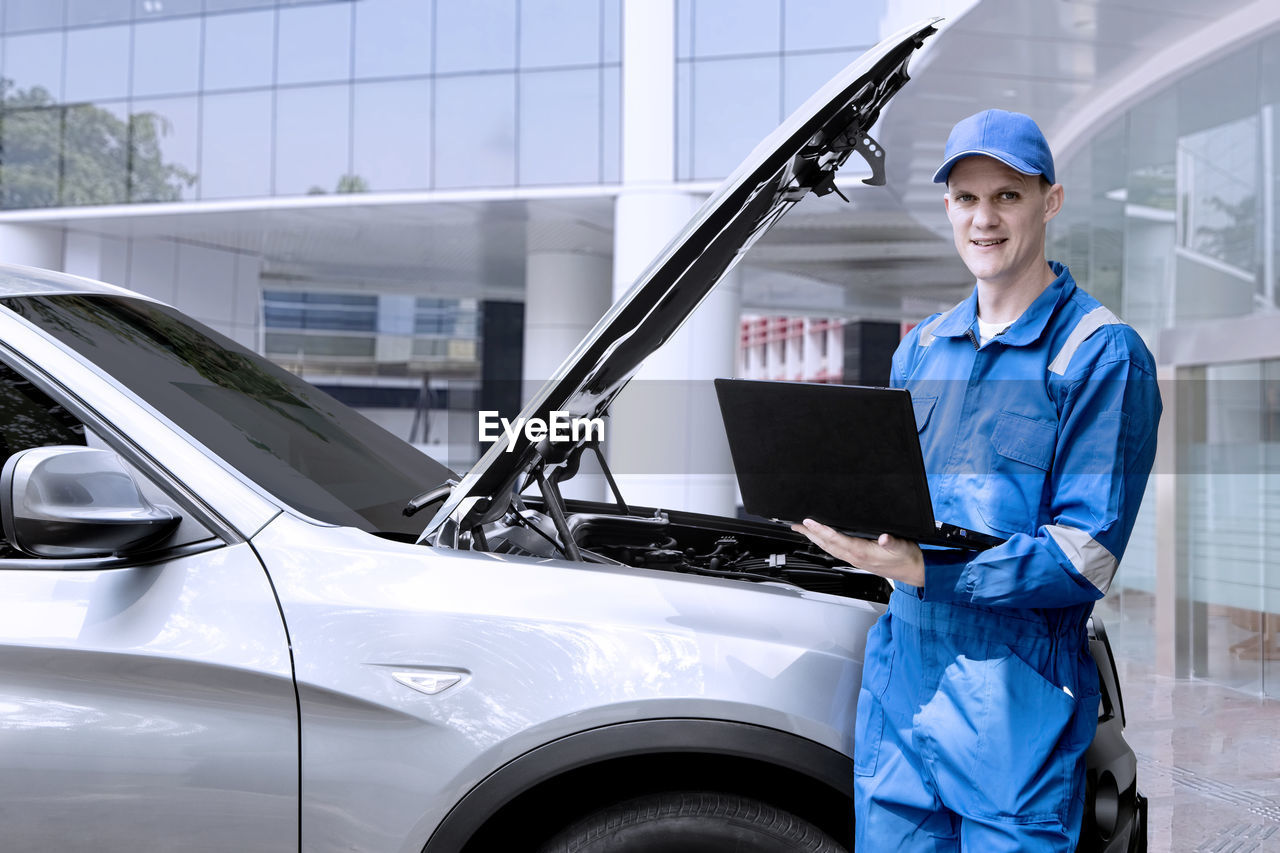 Portrait of smiling mechanic using laptop while standing against car