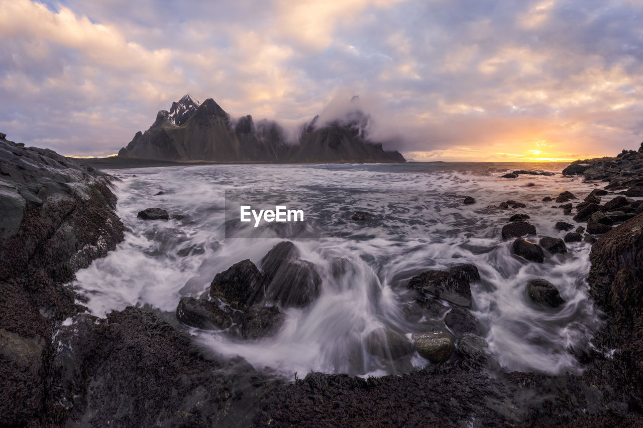 Picturesque scenery of waving sea washing rough shore near high snowy mountains reaching cloudy sky