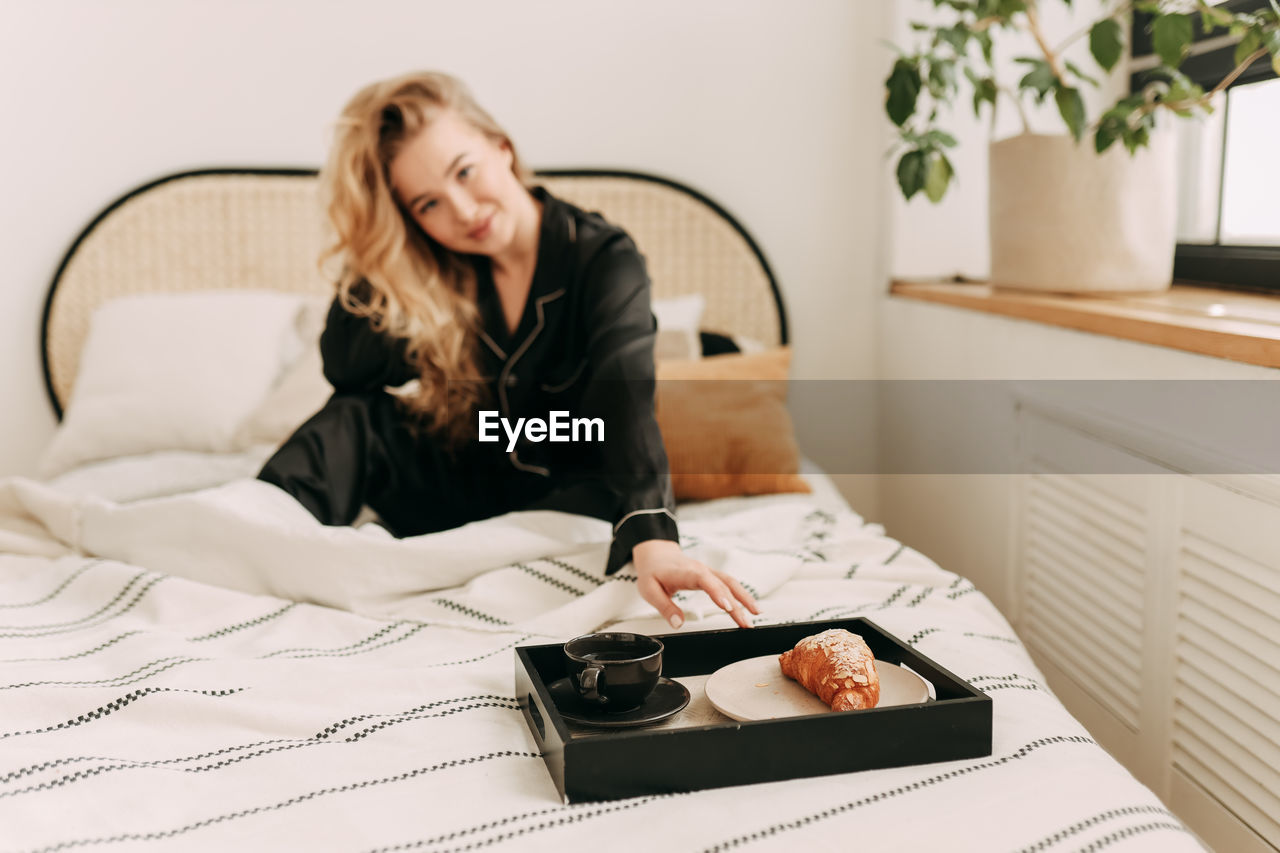 A happy pretty young woman in pajamas is resting and having breakfast in bed in a cozy apartment