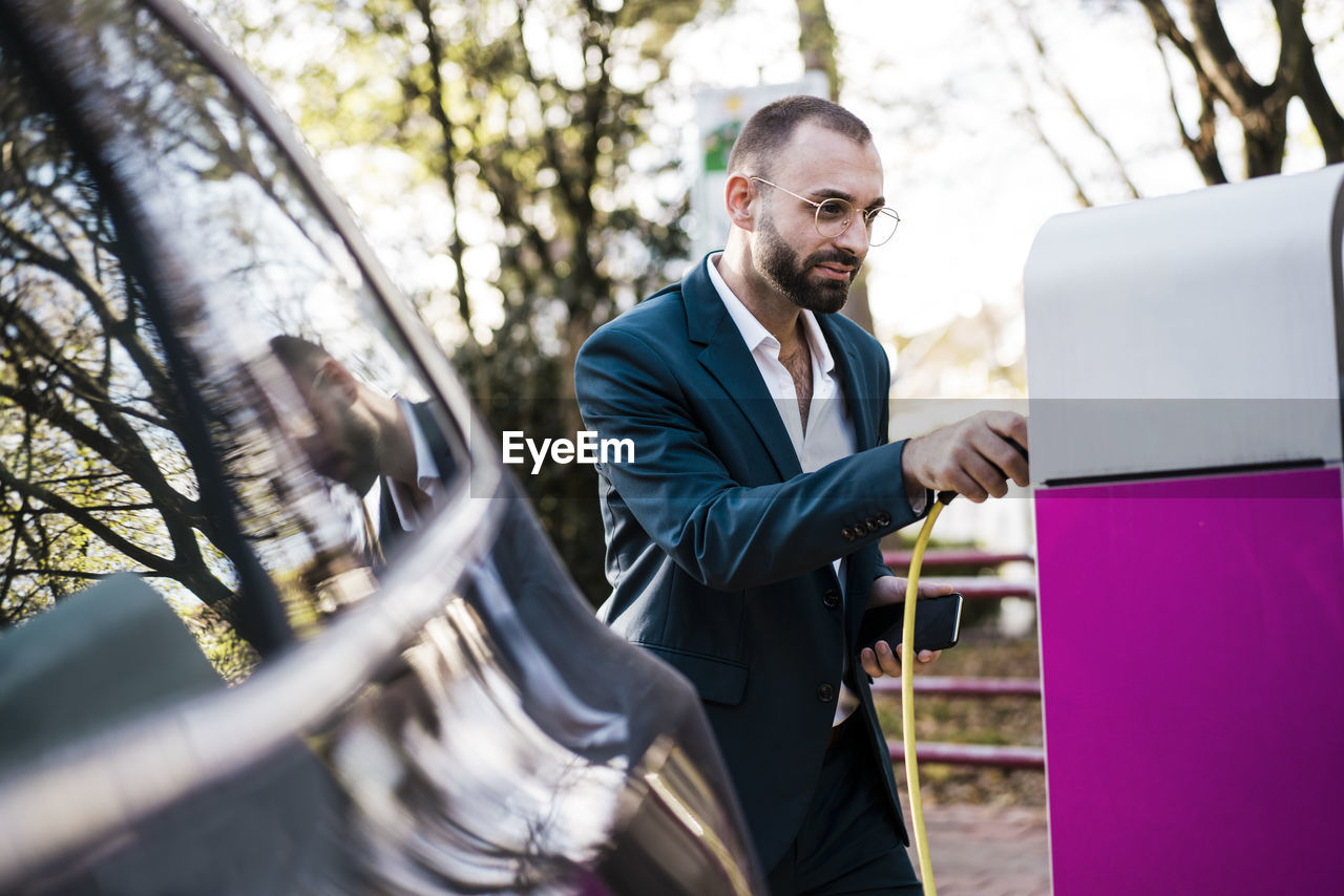 Smiling businessman holding mobile phone at electric vehicle charging station