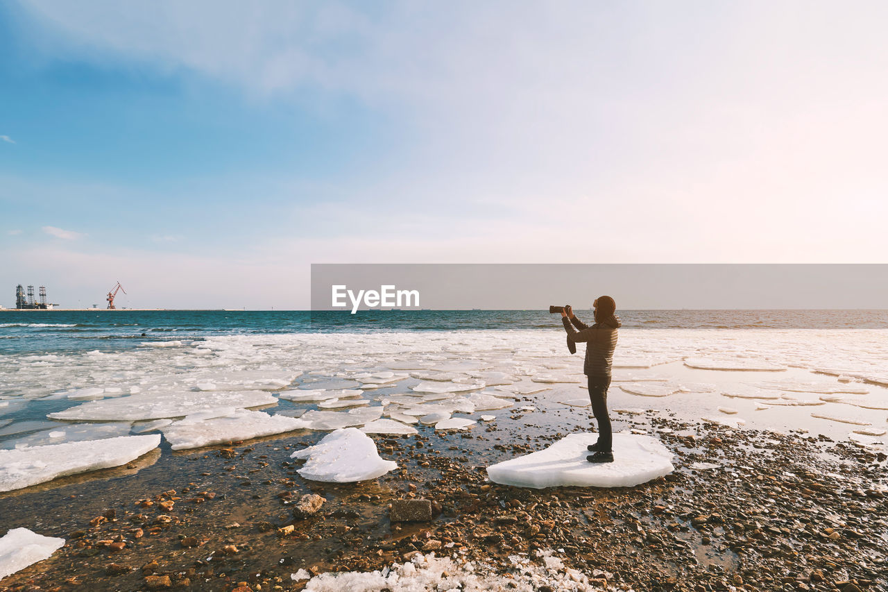 Full length of man photographing through camera while standing on ice against sea at beach