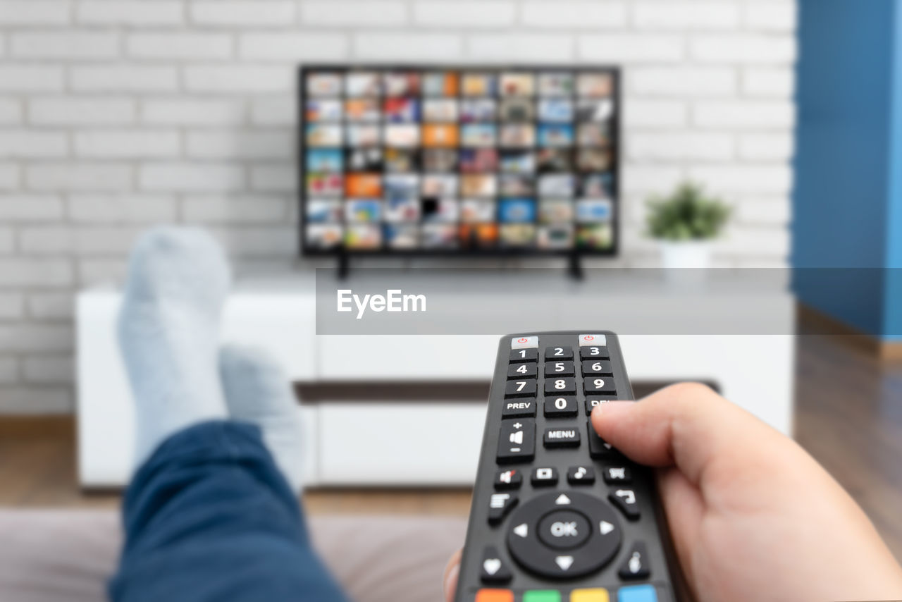 remote control, television set, technology, relaxation, one person, watching tv, hand, indoors, remote, adult, leisure activity, control, living room, men, holding, lifestyles, arts culture and entertainment, domestic room, furniture, home interior, close-up, communication, focus on foreground, gadget