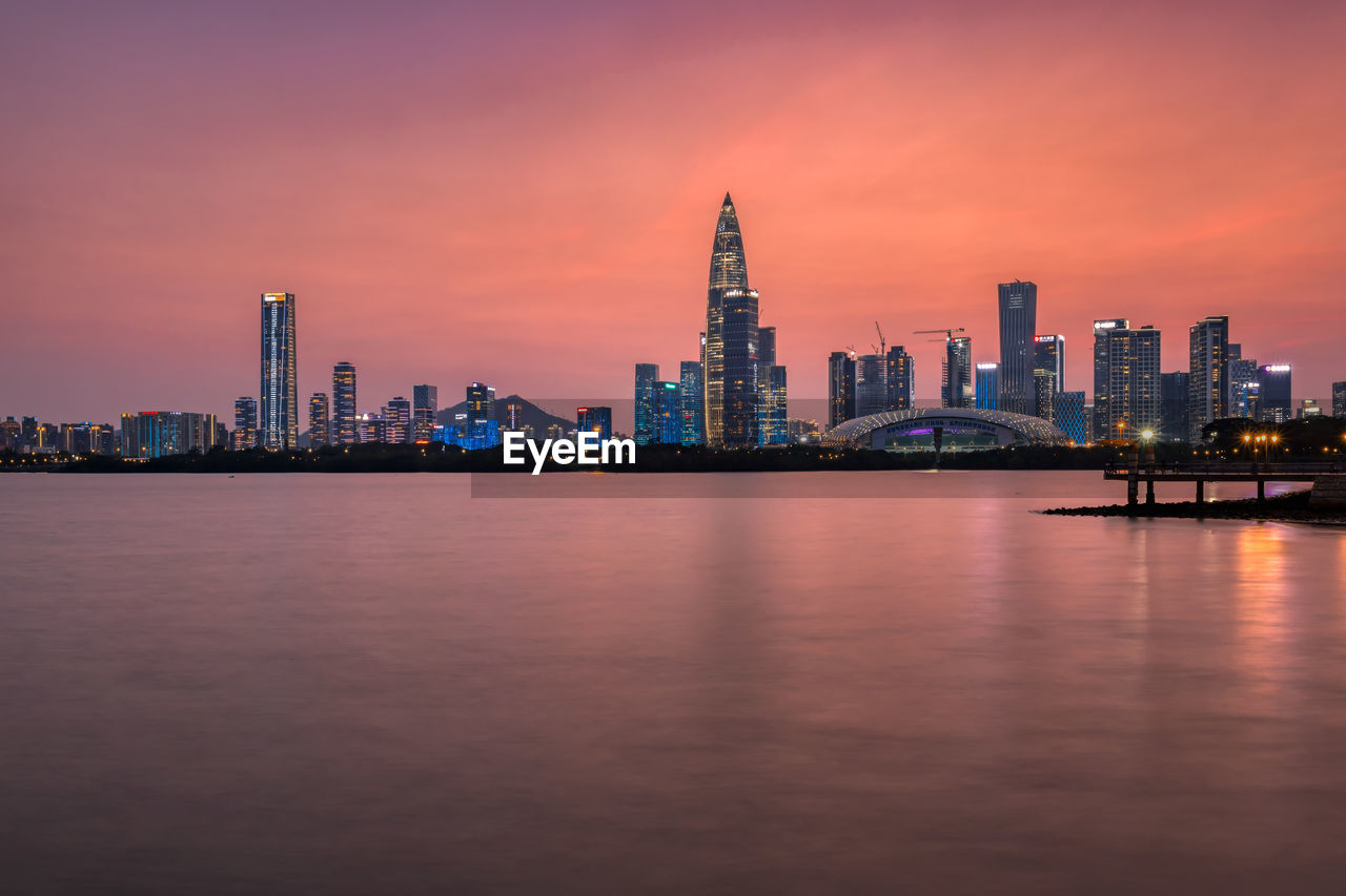 SEA BY ILLUMINATED BUILDINGS AGAINST SKY AT SUNSET