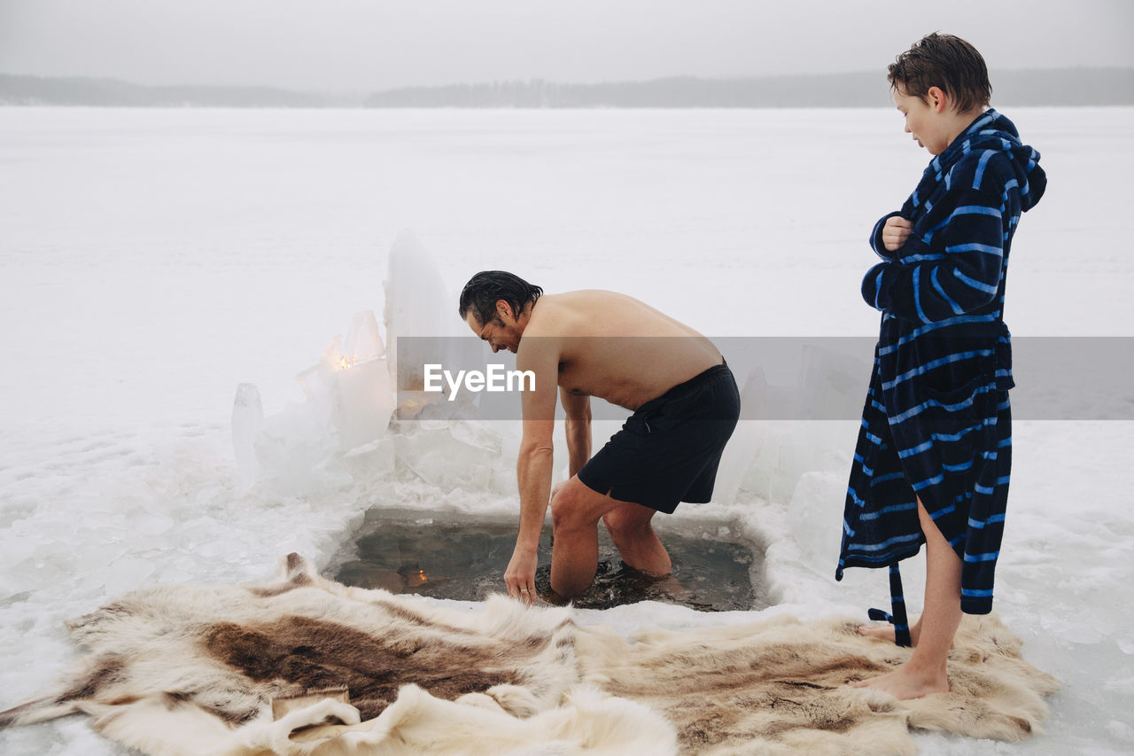 Boy looking at father taking ice bath at frozen lake