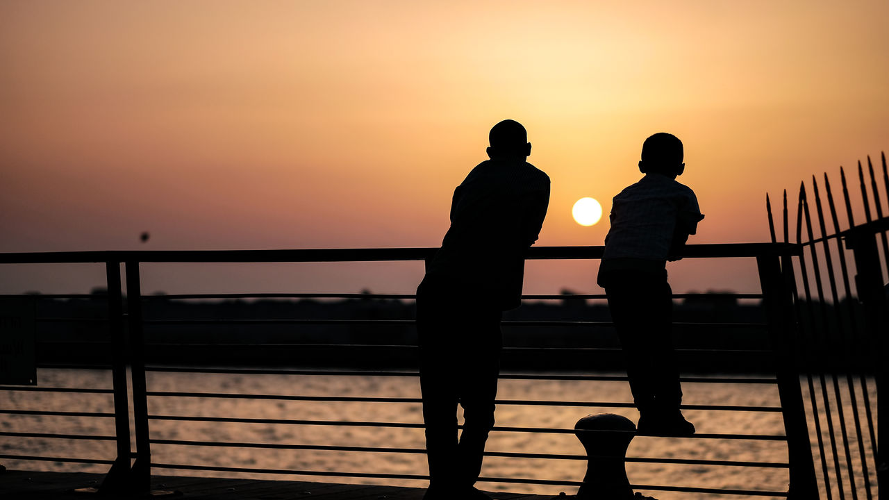 SILHOUETTE PEOPLE STANDING BY RAILING AGAINST ORANGE SKY